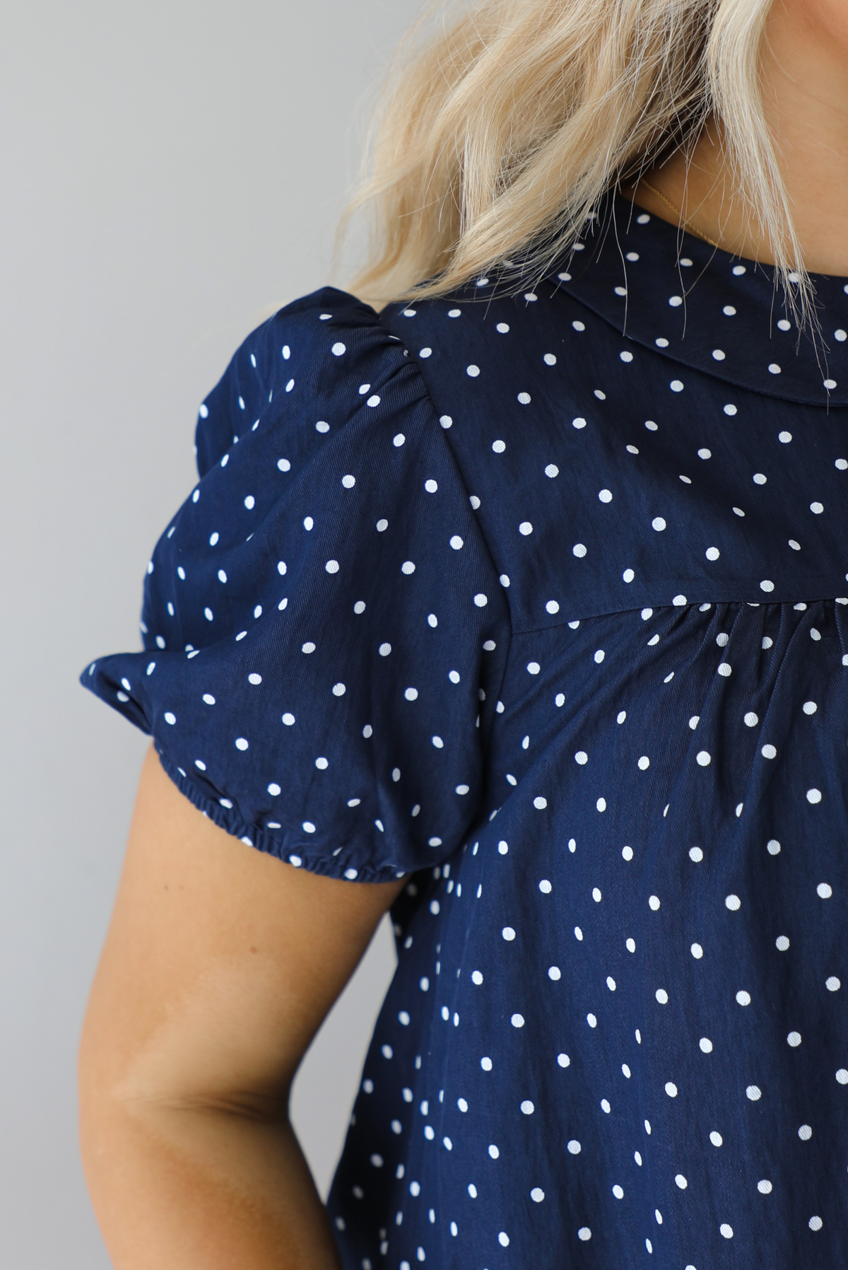 Woman wearing a navy baby-doll cut dress with a white polkadot print pattern throughout. The dress has puffed short sleeves and functional pockets on either side. The photo shows a closeup of the short puffed sleeve that has elastic at the hem. The photo is taken indoors against a white wall.