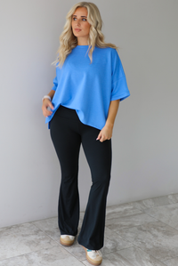 Woman wearing an oversized blue box tee with black leggings that flare slightly at the hem. The photo is taken indoors against a white wall with white marble flooring. 