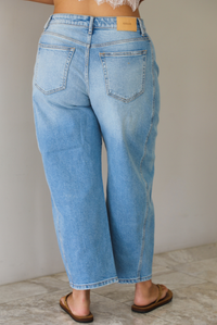 A woman wearing light-wash blue cropped barrel cut jeans with an asymmetrical button closure, standing indoors against white wall with white marble flooring. The photo shows the back of the jeans, which have functional back pockets. 