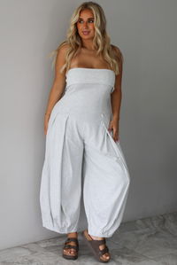A woman modeling a light gray strapless jumpsuit with wide, balloon-style legs and gathered detailing, standing indoors against a white wall with white marble flooring. 