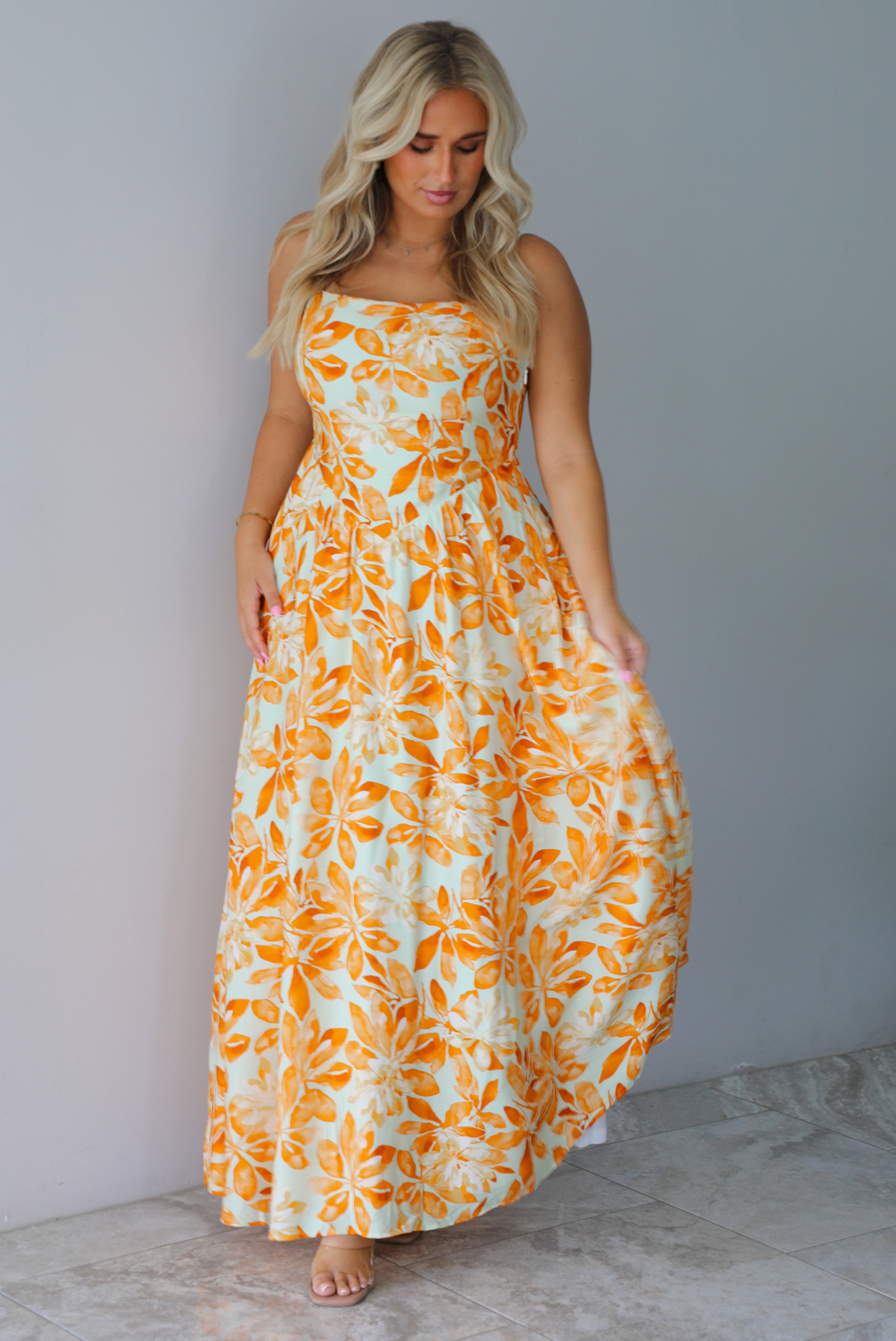 A woman modeling a spaghetti strapped, floor-length maxi dress with a white base and a vibrant marigold orange floral pattern, standing indoors against a white wall with white marble flooring. 
