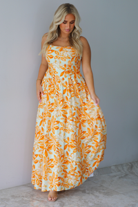 A woman modeling a spaghetti strapped, floor-length maxi dress with a white base and a vibrant marigold orange floral pattern, standing indoors against a white wall with white marble flooring. 