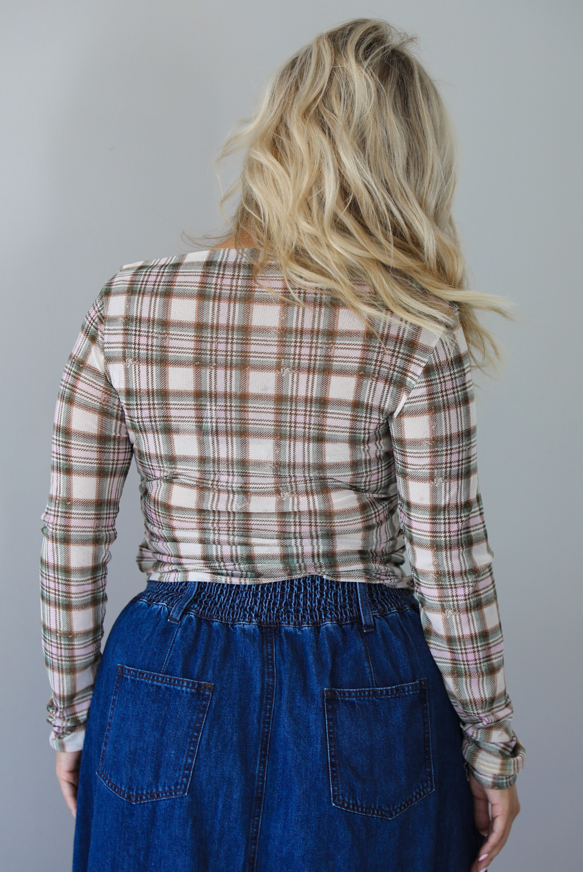 A woman shown from behind modeling an off-the-shoulder long-sleeve plaid top in beige, brown, and green tones, paired with a blue denim skirt, indoors against a white wall. 