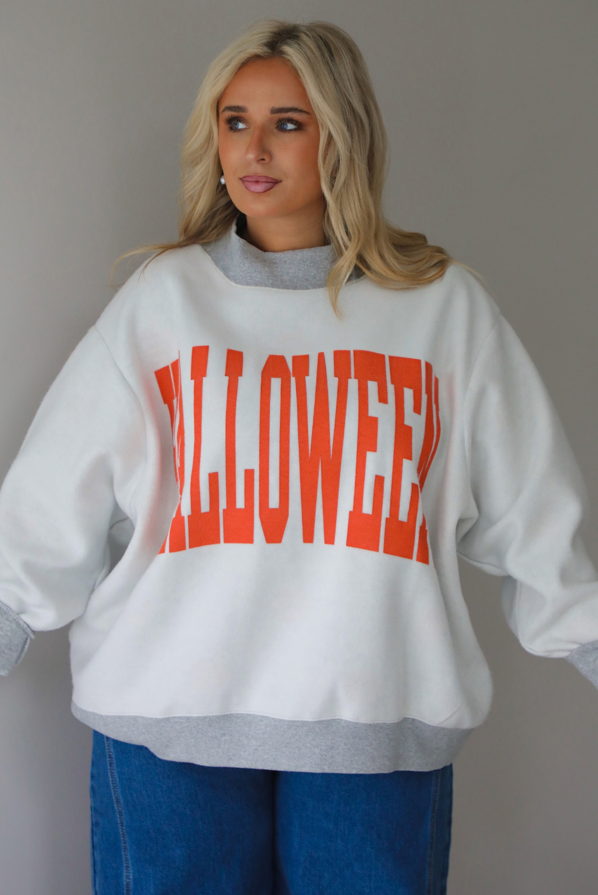 A woman wearing a light grey and dark grey trim oversized sweatshirt that says "HALLOWEEN" in collegiate font on the front, with jack-o-lantern details on the rolled up sleeves, paired with dark wash denim jeans, standing indoors against a white wall.
