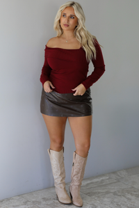 Woman wearing a maroon knit, long sleeved, off the shoulder knit top paired with a dark brown vegan leather mini skirt with a monochromatic polka dot print pattern, and beige knee high boots, standing indoors against a white wall with white marble flooring. 
