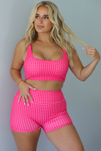 A woman wearing a red and hot pink gingham two-piece activewear set consisting of a crop top and matching shorts, standing indoors against a white wall.