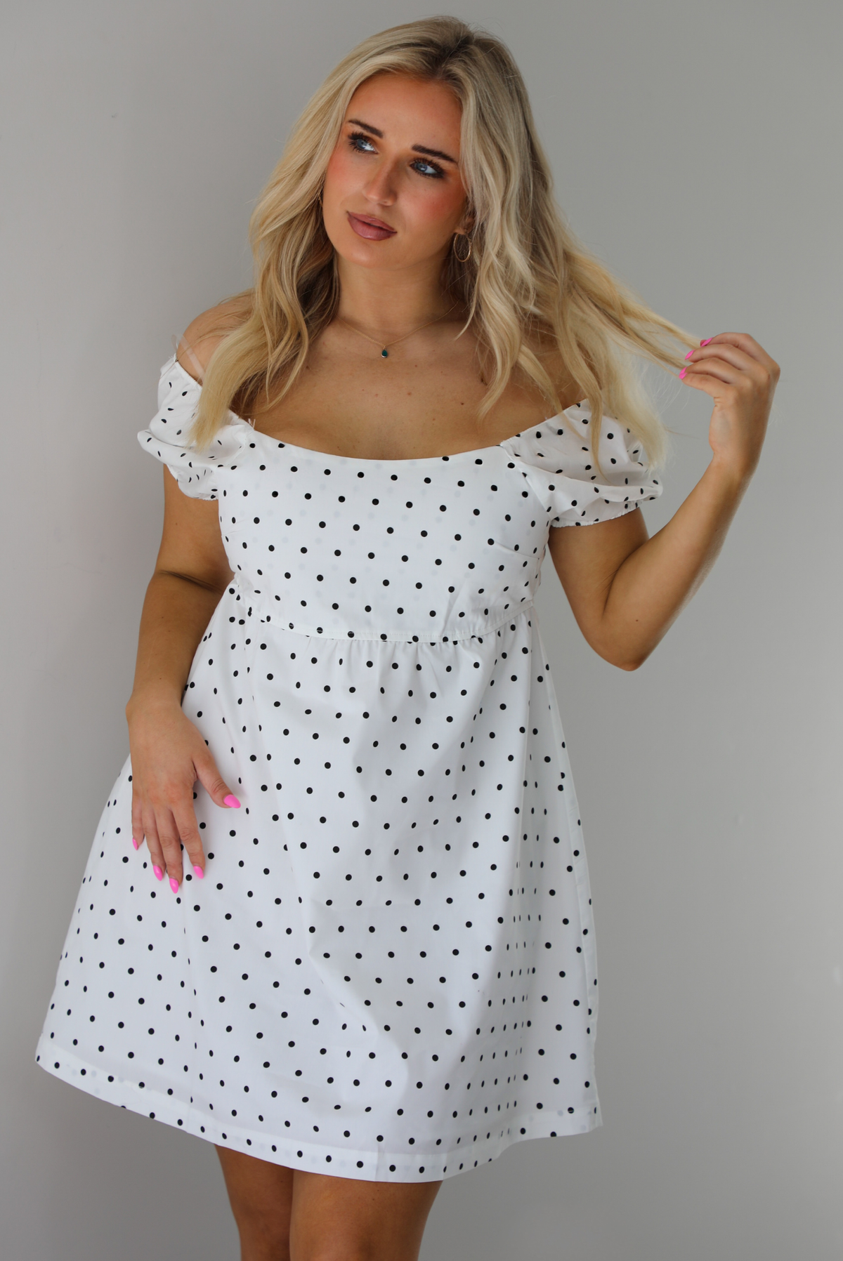Woman wearing a white dress with a black polka dot print throughout. The photo is taken indoors against a white wall.