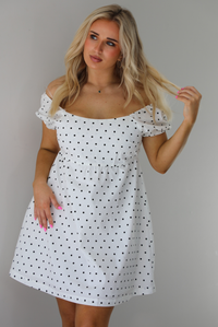 Woman wearing a white dress with a black polka dot print throughout. The photo is taken indoors against a white wall.