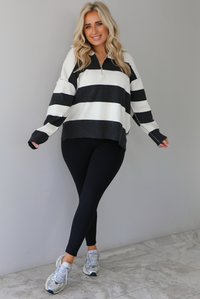 Woman wearing a charcoal and white striped quarter-zip sweater with a collared neckline, raw seam details, and a relaxed fit. It's paired with a fitted black jumpsuit and silver sneakers. The photo is taken indoors against a white wall with white marble flooring.