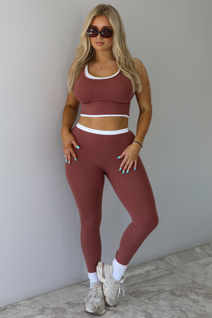Woman wearing a maroon and white layered look two piece athleisure set with a cropped top and high waisted leggings. The photo is taken indoors against a white wall with white marble flooring. 