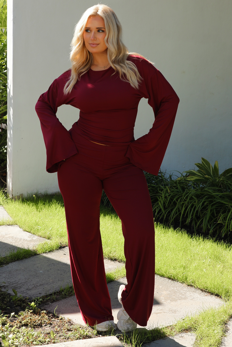 Woman model wearing a burgundy wine colored two piece set outdoors. The set features a slightly cropped, long sleeved top that has flared sleeves and a boat neckline. The pants are a mid-rise, fitted on the thighs, and have a straight cut design at the bottom. It's super soft, lightweight, and stretchy. 