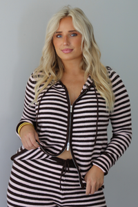A woman wearing a light pink and brown striped two piece set featuring a double zipped hoodie top and mid-rise, wide leg pants, standing indoors against a white wall. The photo shows a closeup of the top and how the top can be zipped from the top or bottom. 