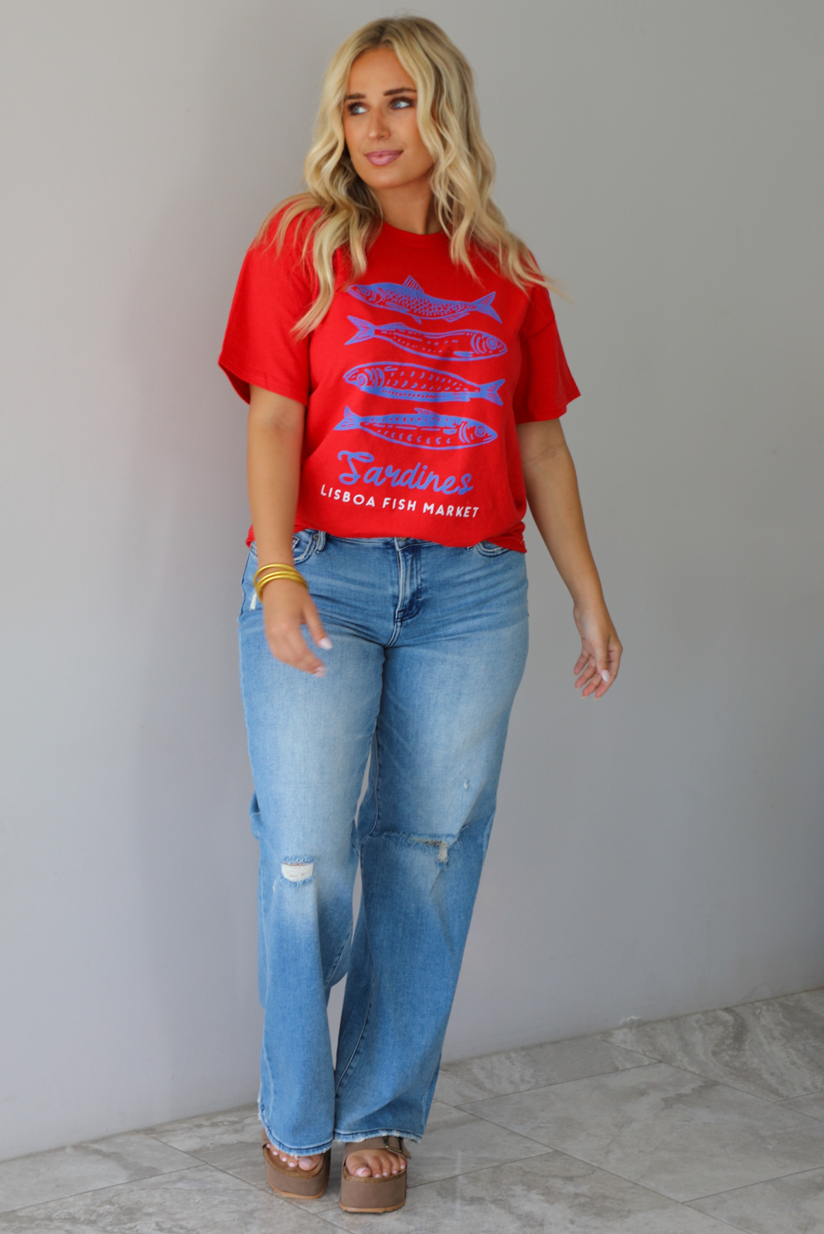A woman wearing a red graphic t-shirt with blue sardine illustrations and the text 'Sardines LISBON FISH MARKET', paired with blue jeans, standing indoors against a white wall with white marble flooring.