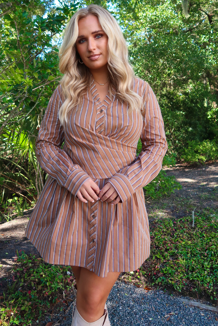 Woman model wearing a rust colored dress that has a black and white vertical striped pattern throughout. The dress has button closures along the front, a wrap detail at the waistline, long sleeves, and a flowy skirt. It is paired with sand colored tall boots that stop just under the knee. The photo is taken outdoors with greenery in the back.
