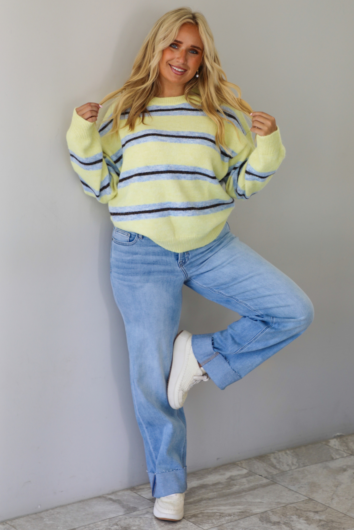 Woman wearing a pastel yellow, blue, and dark brown striped sweater, paired with light wash denim jeans that have a wide leg cut with a cuffed hem, standing indoors against a white wall with white marble flooring.