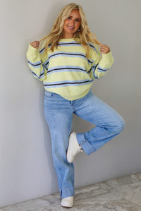 Woman wearing a pastel yellow, blue, and dark brown striped sweater, paired with light wash denim jeans that have a wide leg cut with a cuffed hem, standing indoors against a white wall with white marble flooring.