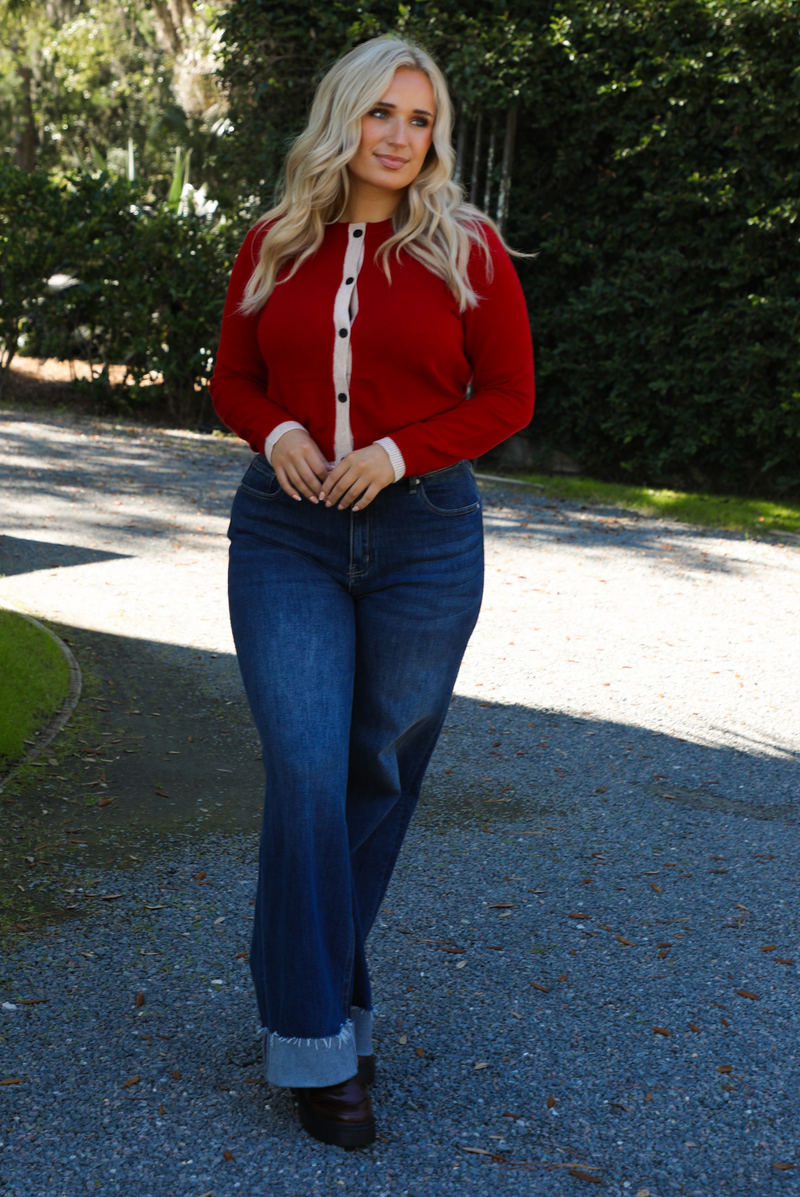 Woman model wearing a red cardigan with sand trim details and black button closures in the front with dark wash denim. The photo is taken outdoors on gravel with greenery in the back. 