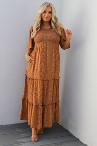 A woman model outdoors wearing a rust dress with a cream floral print pattern throughout. It has flowy short sleeves, a smocked bodice, and a flowy maxi length skirt with functional pockets on either side. 