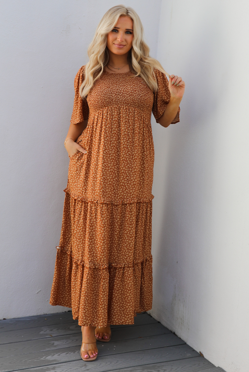A woman model outdoors wearing a rust dress with a cream floral print pattern throughout. It has flowy short sleeves, a smocked bodice, and a flowy maxi length skirt with functional pockets on either side. 