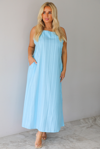A woman modeling a light blue sleeveless maxi dress with vertical grey pinstripes and side pockets, standing indoors against a white wall with white marble flooring. 