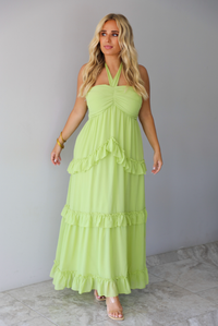 A woman modeling a lime green halter-neck maxi dress with tiered ruffle details, standing indoors against a white wall with white marble flooring. 