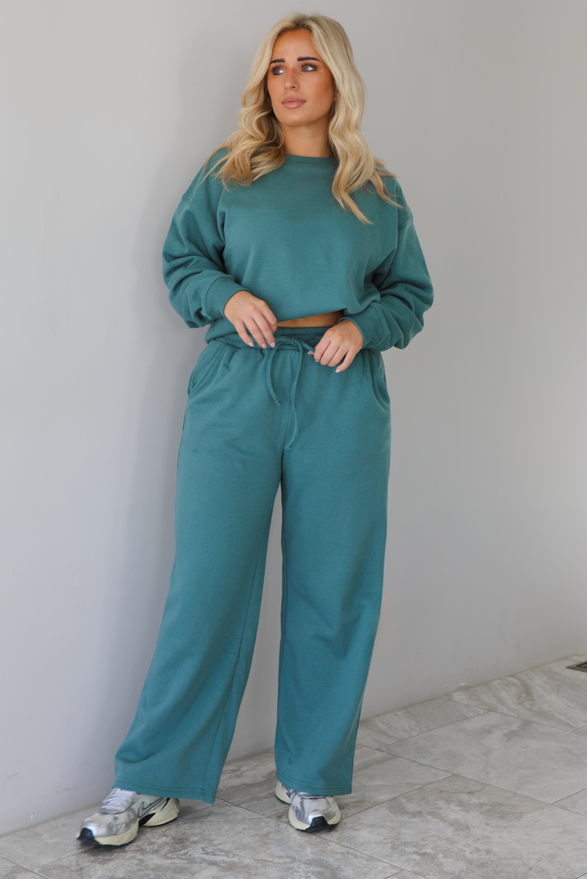 Woman wearing a sage two piece sweat-set. The set features a slightly oversized sweater and high waisted sweat pants that have an elastic and drawstring waistline with pockets. The photo is taken indoors against a white wall with white marble flooring. 