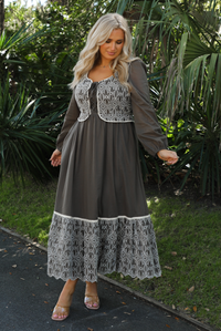 Woman model wearing an olive and off-white stitched detail dress outdoors. Dress has a corset-appearance bodice, long sleeves, functional pockets on either side, an elastic waistline, and a long, maxi length.