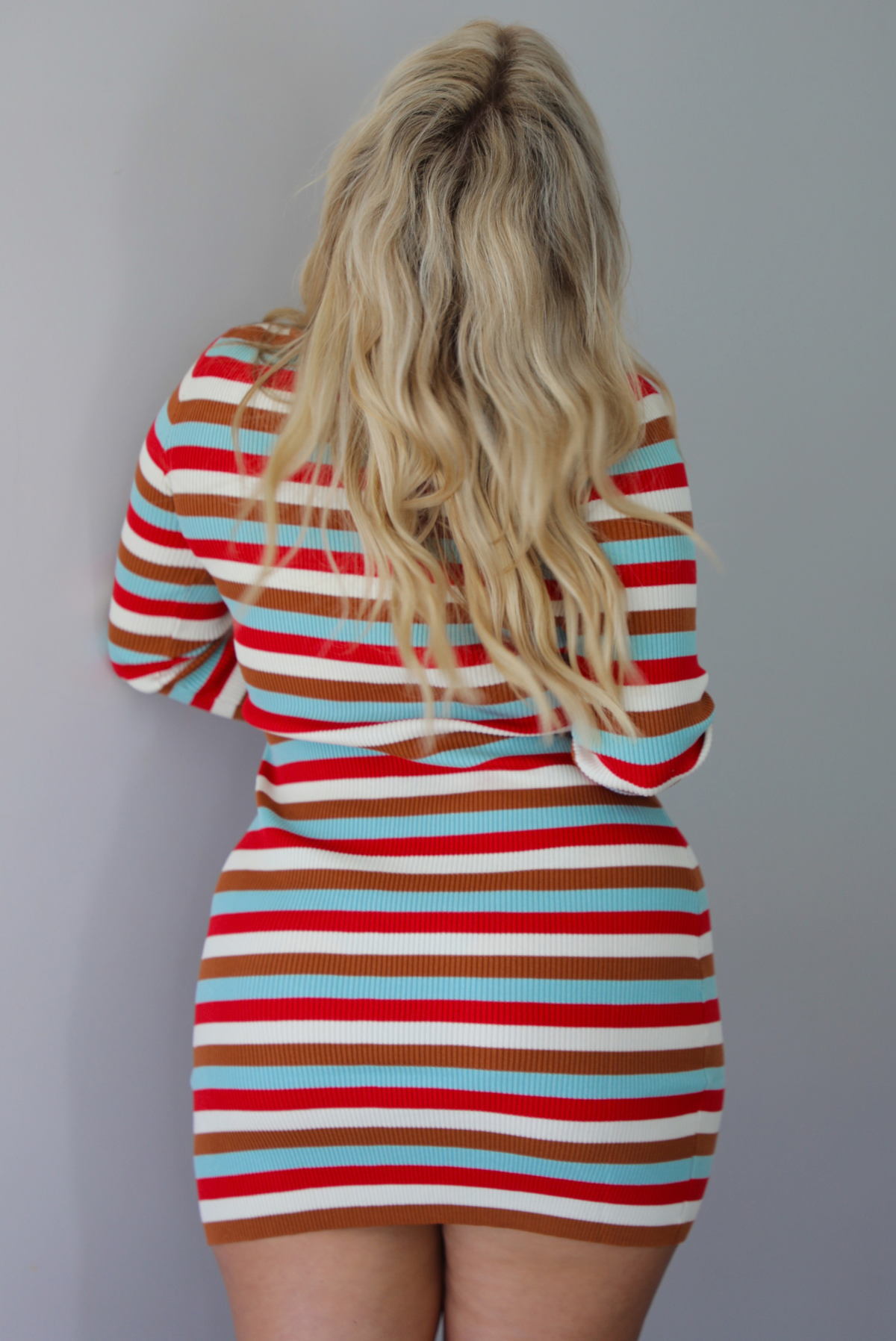 A woman shown from behind wearing a red, blue, white, and brown striped long sleeved mini dress, standing indoors against a white wall.