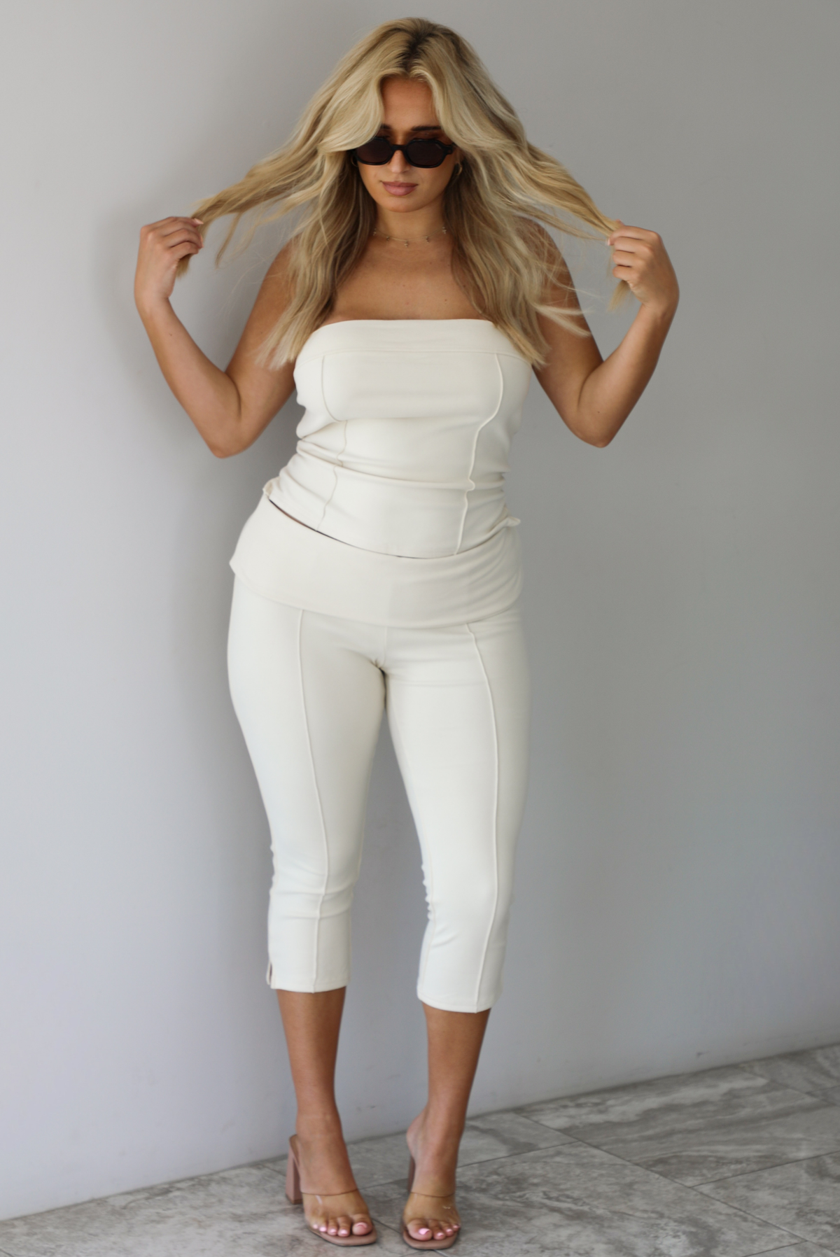 Woman wearing a cream two piece set featuring a strapless tube top and capri pants that fold over at the waist indoors against a white wall with white marble flooring. 