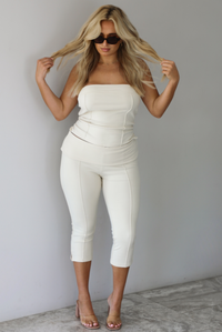 Woman wearing a cream two piece set featuring a strapless tube top and capri pants that fold over at the waist indoors against a white wall with white marble flooring. 