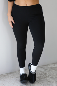 Woman wearing a black two piece set featuring an oversized t-shirt and fitted leggings. The photo shows a closeup of the leggings. The outfit is paired with white ankle socks and black slip on shoes. The photo is taken indoors against a white wall with white marble flooring.