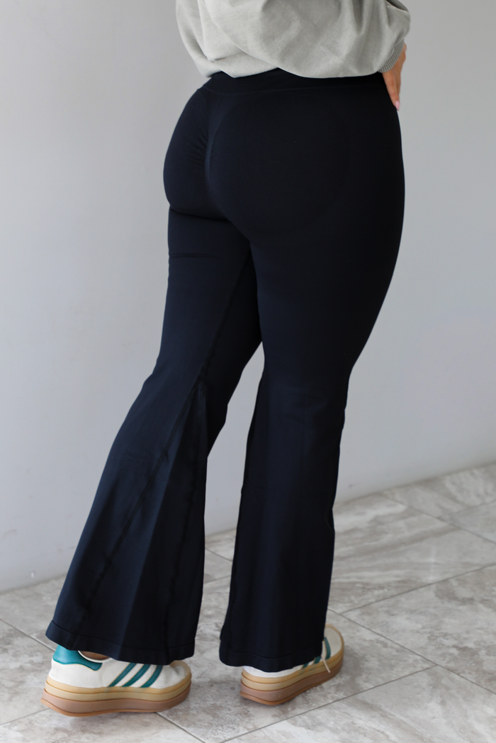 Woman wearing black leggings that are high waisted, fitted, with a flared hem with split hem details. The photo shows the back of the leggings and the scrunched detail at the bottom. The photo is taken in front of a white wall with white marble flooring. 