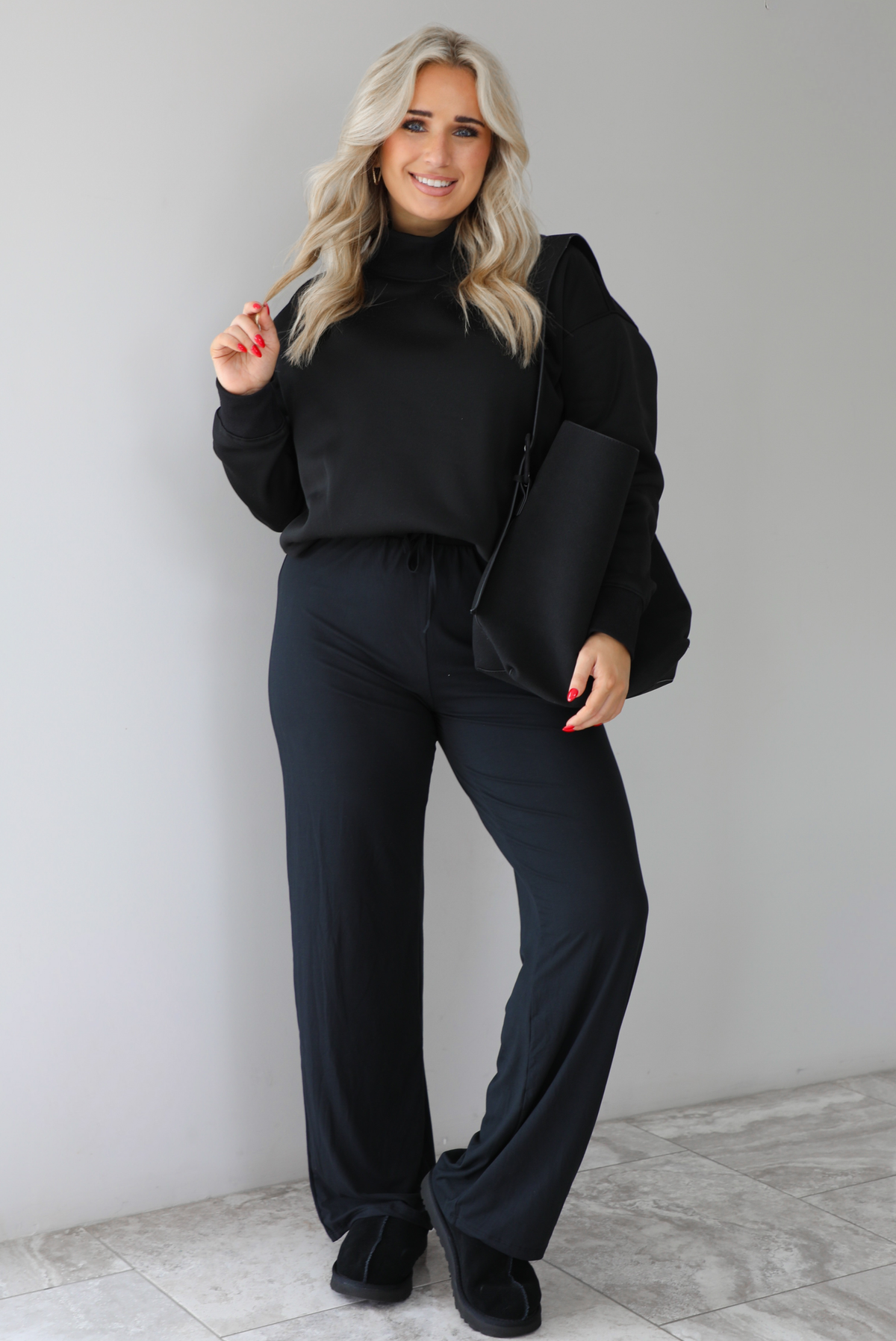 Woman wearing a black high-neck, oversized sweater, high waisted black pants, with a black shoulder bag, and black ugg boot shoes. The photo is taken indoors against a white wall with white marbled flooring. 