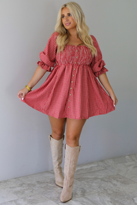 A woman wearing a red and white gingham mini dress with puff, elbow length sleeves and button detailing along the front, paired with beige knee-high boots, standing indoors against a white wall with white marble flooring. 