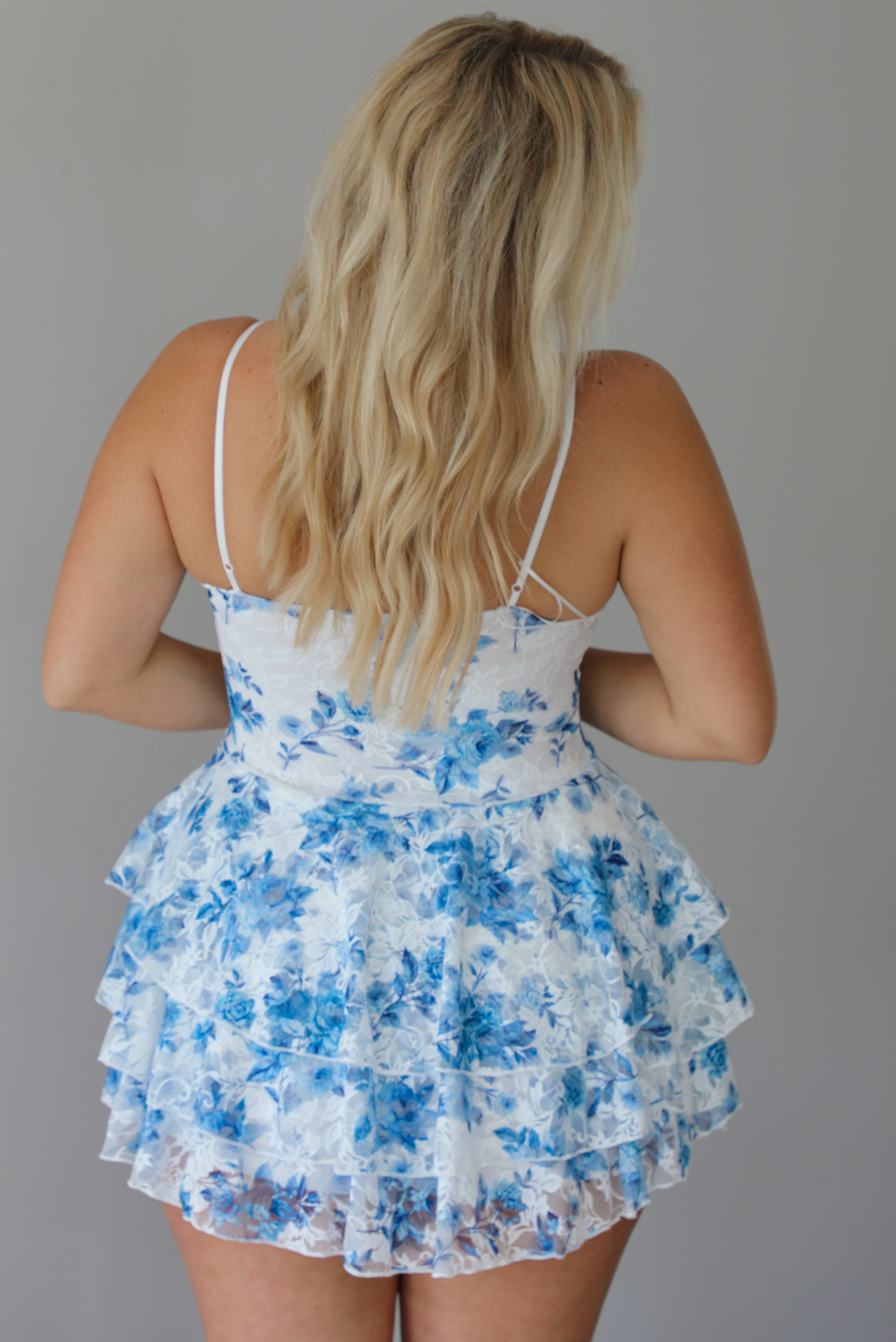 Woman wearing a white and blue floral mini dress with a low neckline, fitted bodice, and flowy, ruffled skirt indoors against a white wall. The photo shows the back of the dress.