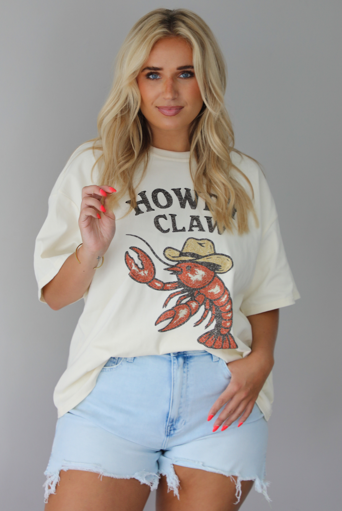Woman wearing a graphic t-shirt with a lobster wearing a cowboy hat and the words 'HOWDY CLAW' written along the front and denim shorts against a white wall. The photo shows the tee front tucked into the denim shorts. 