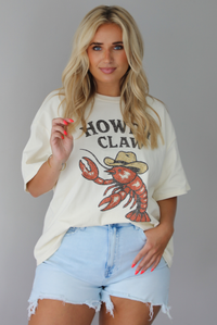 Woman wearing a graphic t-shirt with a lobster wearing a cowboy hat and the words 'HOWDY CLAW' written along the front and denim shorts against a white wall. The photo shows the tee front tucked into the denim shorts. 