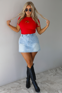 Woman wearing a red fitted baby tee with an embroidered horse graphic on the front. The top is paired with a high waisted, light-wash denim skort. The photo is taken indoors against a white wall with white marble flooring. 