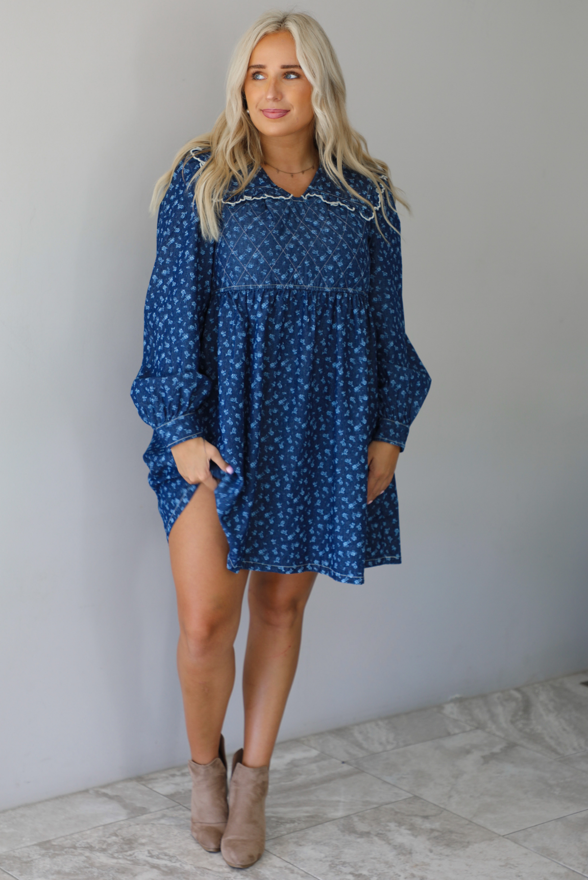 A woman wearing a dark denim mini dress with a white floral print pattern throughout, collared neckline, a quilted detail at the bodice, long sleeves, and a flowy skirt, standing indoors against a white wall with white marble flooring. 