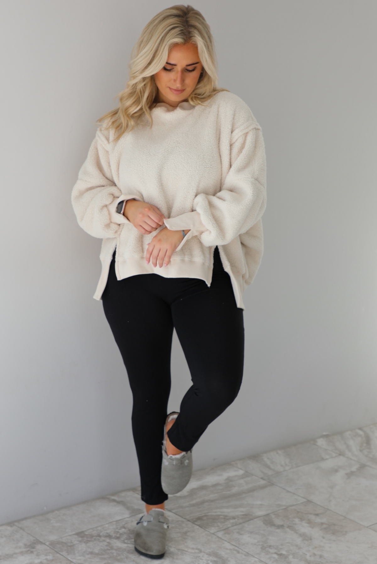 Woman wearing a sherpa sand oversized sweater with a fitted black jumpsuit underneath and grey slide on shoes. The photo is taken indoors against a white wall with white marble flooring. 