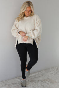 Woman wearing a sherpa sand oversized sweater with a fitted black jumpsuit underneath and grey slide on shoes. The photo is taken indoors against a white wall with white marble flooring. 