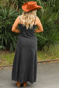 Woman wearing a black denim maxi length dress that has thin, adjustable spaghetti straps, cup stitching at the bust with other stitched details, slight distressing at the thighs, functional button closures in the front, with a heavier weight material with some stretch throughout, smocked material in the back, and is not lined. The photo shows the back of the dress with its smocked material.