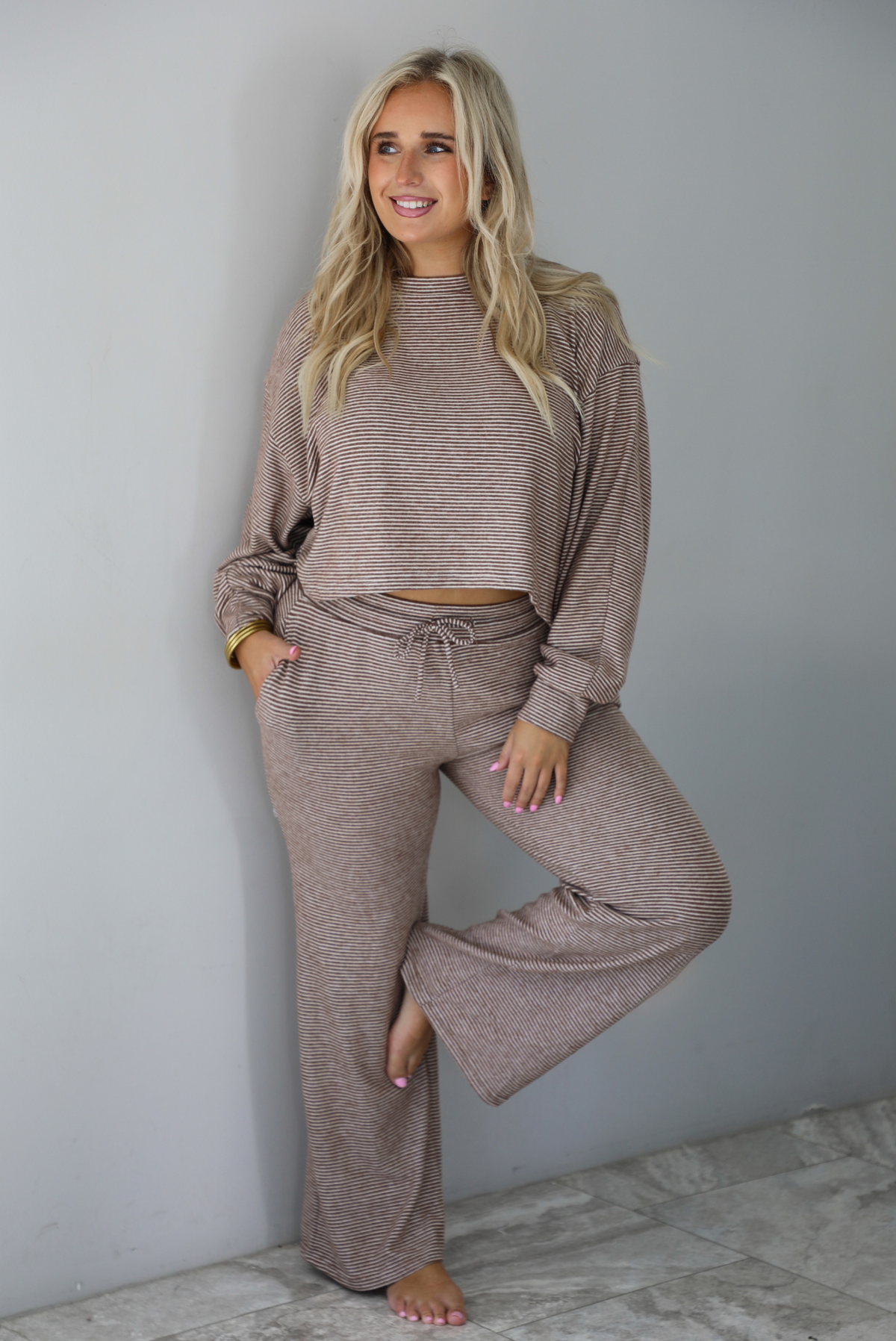 A woman wearing a mocha and white striped matching two piece set featuring a cropped, flowy. long sleeved top paired with high waisted wide leg pants, standing indoors against a white wall with white marble flooring. 