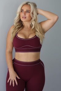 A woman wearing a maroon matching two piece set featuring a sports bra and high waisted leggings with white stitching details, standing indoors against a white wall.