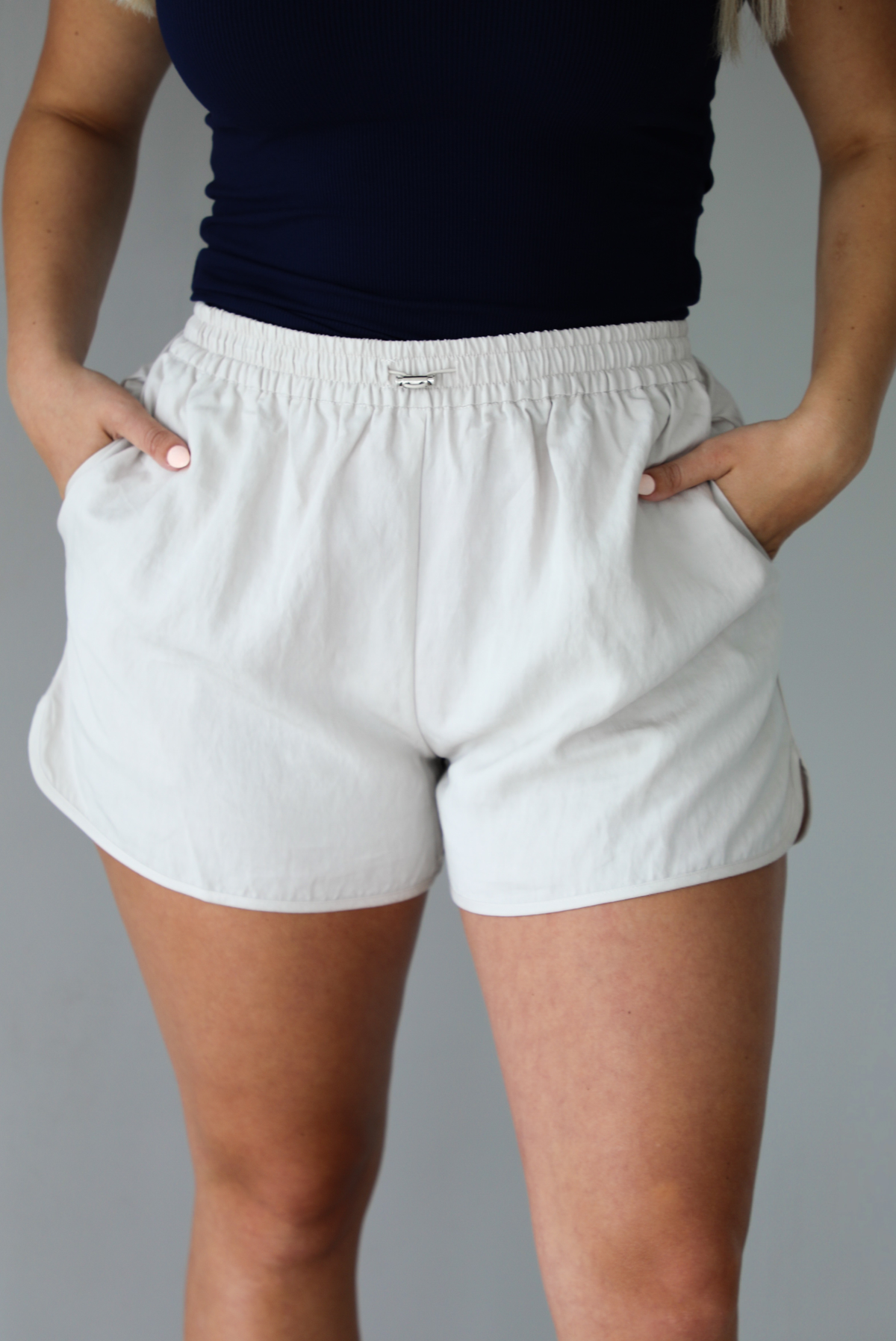 Woman wearing a stone colored two piece set featuring a zip up jacket and shorts with a navy tank tucked into the shorts indoors against a white wall. The photo shows a closeup of the shorts, which have an elastic waistline, drawstring waistline, and pockets. 