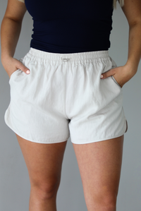 Woman wearing a stone colored two piece set featuring a zip up jacket and shorts with a navy tank tucked into the shorts indoors against a white wall. The photo shows a closeup of the shorts, which have an elastic waistline, drawstring waistline, and pockets. 