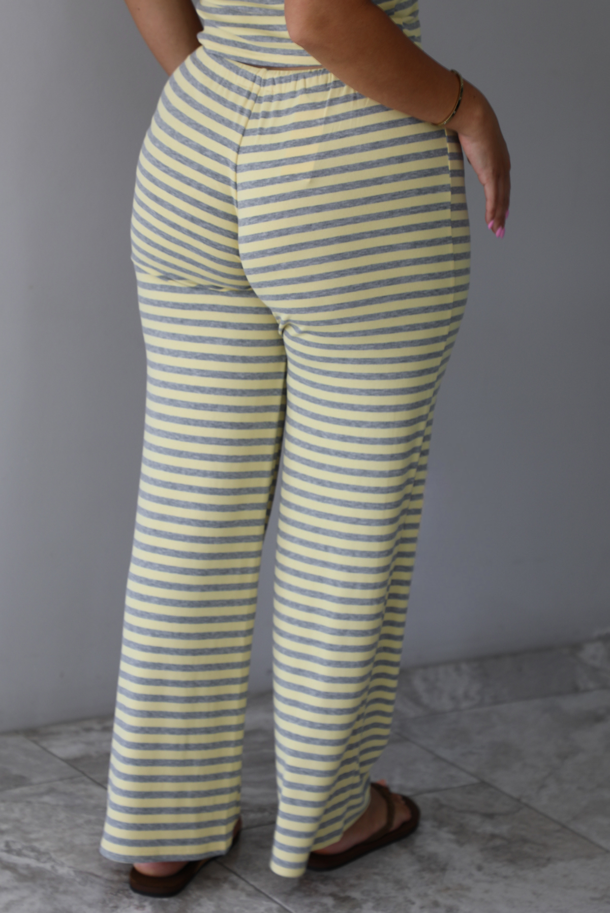 A woman wearing a two piece set consisting of a collared, button-down, short sleeve top and high waisted, drawstring, wide leg pants, standing indoors against a white wall with white marble flooring. It shows a closeup of the back of the pants.