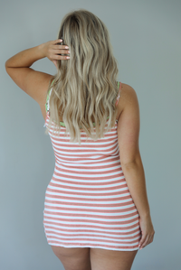 Woman wearing an orange, white, and green striped dress that looks layered with a green dress underneath indoors against a white wall. The photo shows the back of the dress, which shows the adjustable straps. 