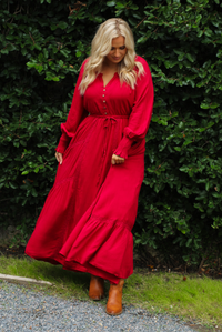 Woman wearing a red maxi length dress that has a v-neckline with button closures at the bodice, a cinched with elastic waistline, long sleeves, and a flowy skirt. The photo is taken outdoors against greenery. 