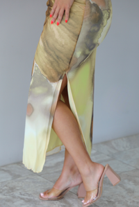 Woman wearing a yellow and brown floral print, strapless, fitted midi dress indoors against a white wall with white marble flooring. The photo shows a closeup of the split hem detail on the left side of the dress. 